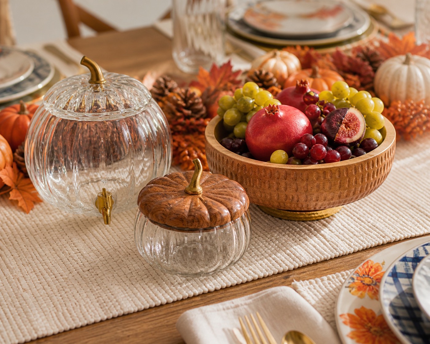 Low fall centerpiece with glass pumpkin cloche, wood bowl of fruit, and pinecones along a table runner